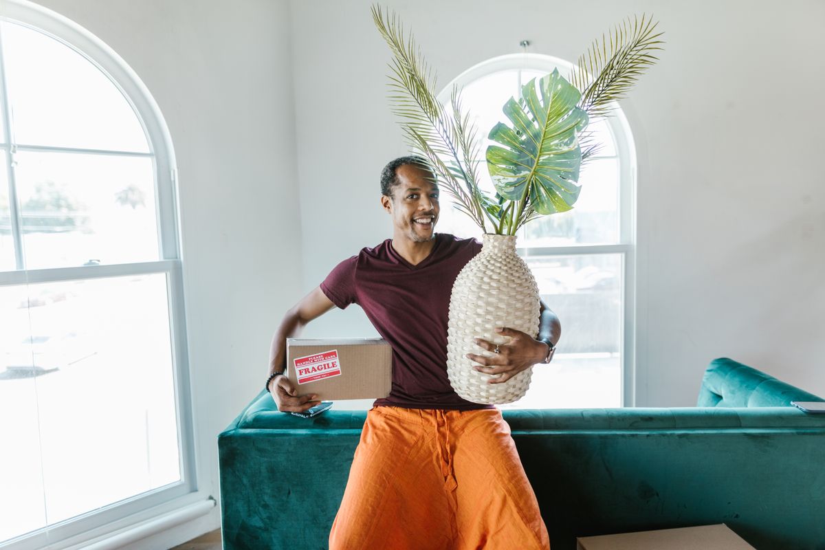A mover carrying a large houseplant and a box into a new home
